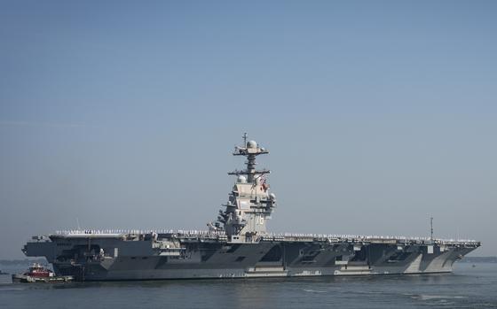 An aircraft carrier, seen from a distance, sails from its berth with a smaller tugboat in front.