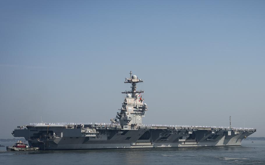An aircraft carrier, seen from a distance, sails from its berth with a smaller tugboat in front.