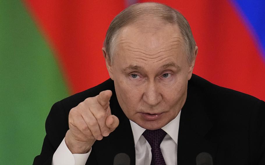 Vladimir Putin raises his right hand and points with his index finger as he leans forward to speak into two microphones.