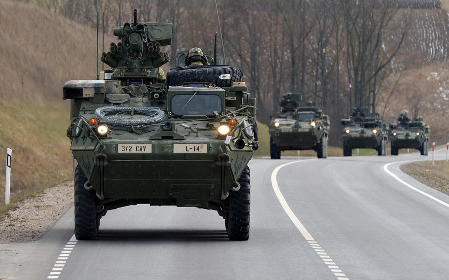 A convoy of armored vehicles moves along a paved road through a countryside. The lead vehicle is a wheeled armored personnel carrier with “3/2 CAV” and “L-14” markings visible on its front. Additional armored vehicles follow behind in formation. 
