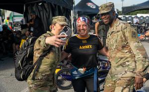 Two U.S. Army soldiers and a female athlete wearing an “ARMY” T-shirt and a patriotic headband smile for a selfie at an outdoor event. The group is surrounded by tents, people, and adaptive sports equipment in the background.