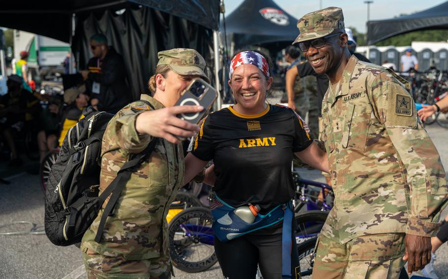 Two U.S. Army soldiers and a female athlete wearing an “ARMY” T-shirt and a patriotic headband smile for a selfie at an outdoor event. The group is surrounded by tents, people, and adaptive sports equipment in the background.