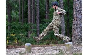 A man in military uniform jumps between raised logs that are far apart.