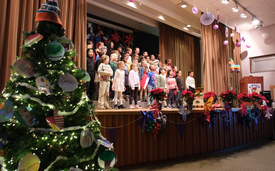 A group of students sing on stage in the center of the photo, while a Christmas tree is seen in the foreground on the left side.