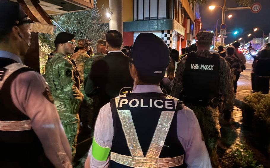 A Japanese police officer in the foreground wears a vest reading “Police” as some U.S. Navy service members stand in the background.