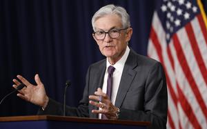 Jerome Powell in a gray suit and purple tie at a podium, both hands raised in gesture as he speaks.