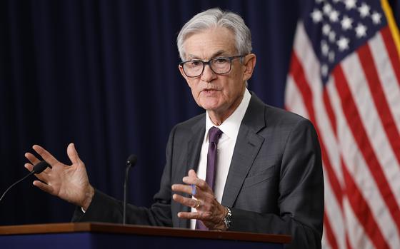Jerome Powell in a gray suit and purple tie at a podium, both hands raised in gesture as he speaks.