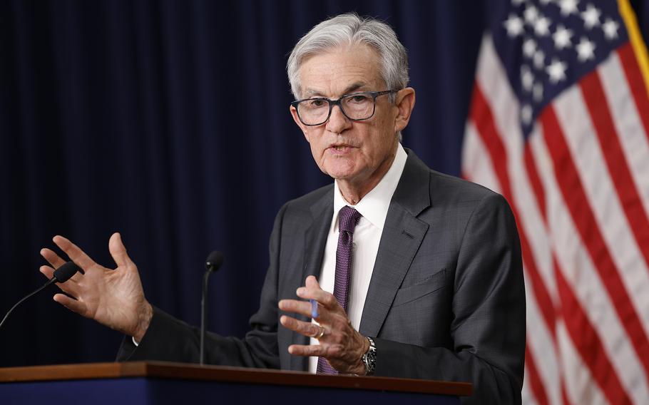Jerome Powell in a gray suit and purple tie at a podium, both hands raised in gesture as he speaks.