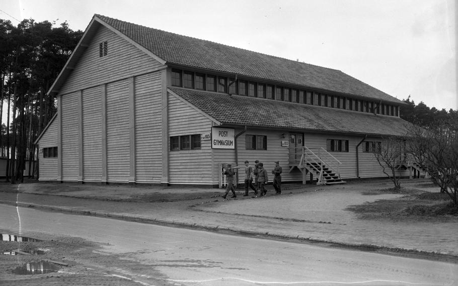 A black and white photo of a long building by a road, with five people walking past the building.