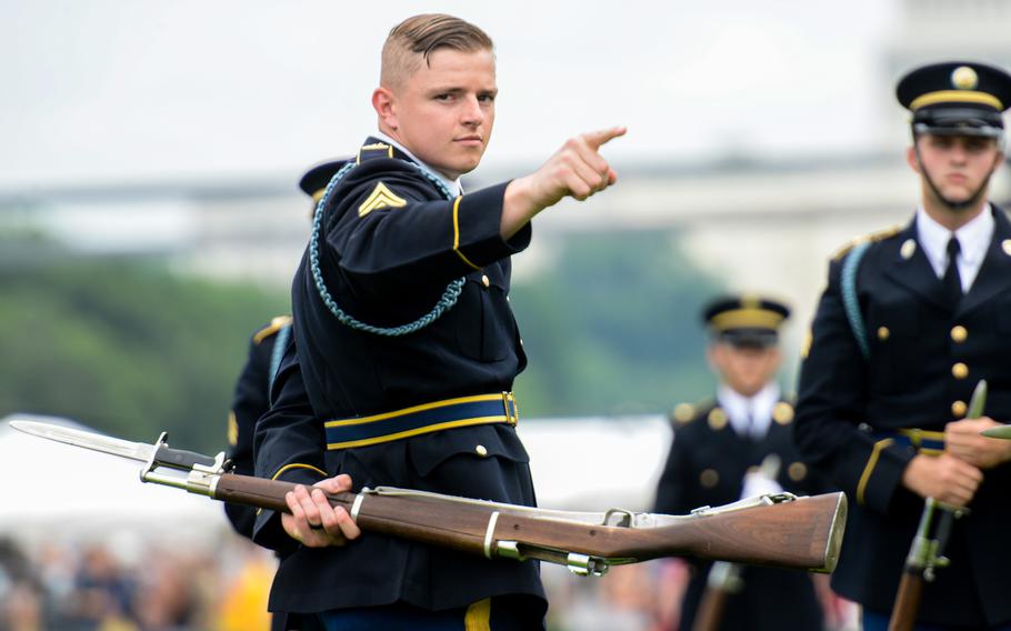 Soldiers with the Army Drill Team on a cloudy day, with one pointing his index finger forward as he holds a rifle behind his back with his other hand.