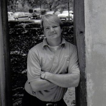 A black-and-whiteimage of a man with his arms folded across his body leaning against a wooden door frame.