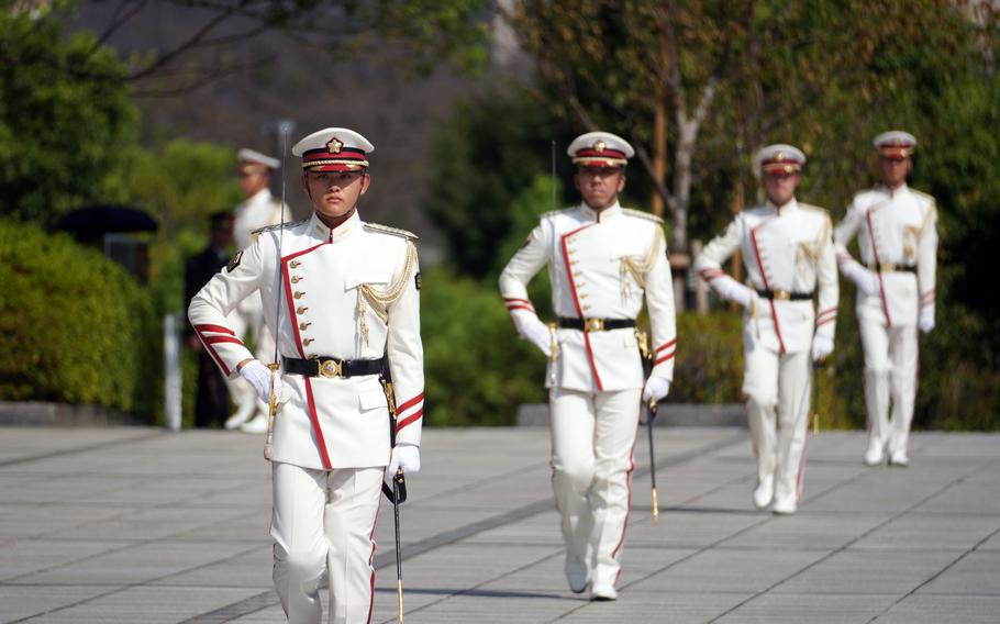 A ceremonial honor guard in formal white military dress uniforms with red trim and white peaked caps marches in formation on a stone courtyard. 