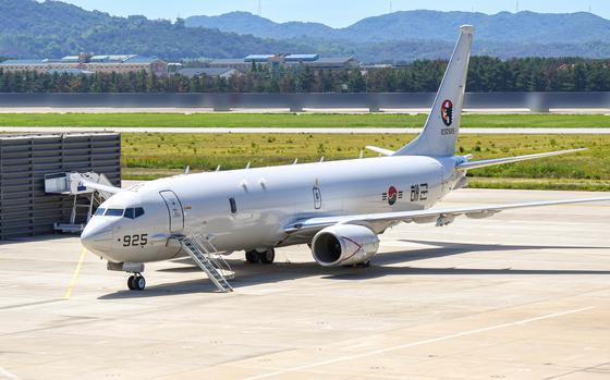 A large military aircraft, seen from a distance, sits parked on a runway, with a treeline and the horizon in the background.