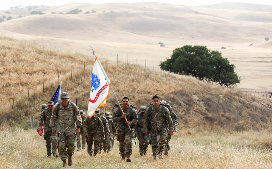A group of soldiers, seen from the front, march up a grassy hill. Two are carrying flags.