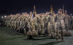 Rows of soldiers in desert camouflage uniforms stand in formation at night. In the foreground, military personnel hold ceremonial flags on poles, with their gear bags placed on the ground in front of them.