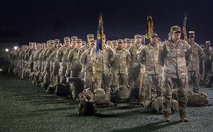 Rows of soldiers in desert camouflage uniforms stand in formation at night. In the foreground, military personnel hold ceremonial flags on poles, with their gear bags placed on the ground in front of them.
