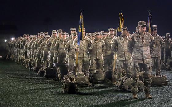 Rows of soldiers in desert camouflage uniforms stand in formation at night. In the foreground, military personnel hold ceremonial flags on poles, with their gear bags placed on the ground in front of them.