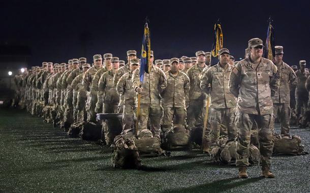Rows of soldiers in desert camouflage uniforms stand in formation at night. In the foreground, military personnel hold ceremonial flags on poles, with their gear bags placed on the ground in front of them.