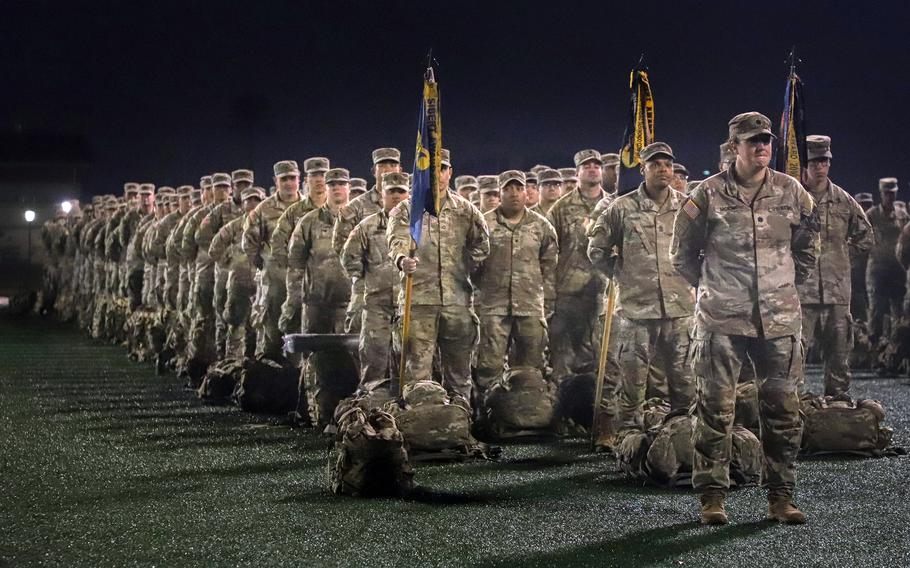 Rows of soldiers in desert camouflage uniforms stand in formation at night. In the foreground, military personnel hold ceremonial flags on poles, with their gear bags placed on the ground in front of them.