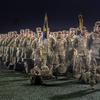 Rows of soldiers in desert camouflage uniforms stand in formation at night. In the foreground, military personnel hold ceremonial flags on poles, with their gear bags placed on the ground in front of them.