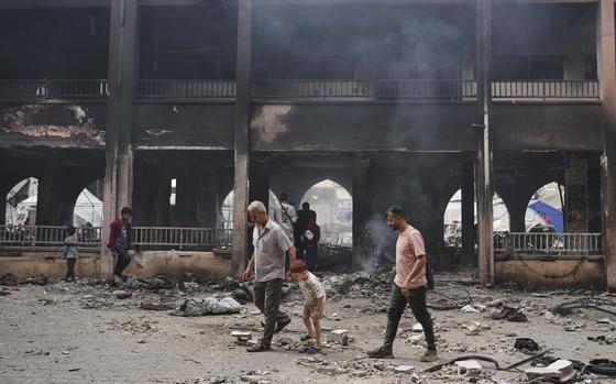 Men and children walk through a rubble-strewn floor before a burned building facade.