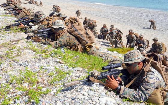 A row of Marines in camouflage combat gear aim rifles while lying on their stomachs atop a dune of rocks and sand, as another row of Marines in camouflage combat gear kneels on a beach behind the with the ocean shoreline in the background.