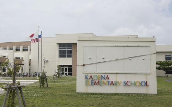 Exterior view of Kadena Elementary School, a modern beige concrete building with large windows and geometric architecture. The school name is displayed in colorful letters on a concrete wall sign in the foreground. Japanese and American flags fly on flagpoles in front of the building.