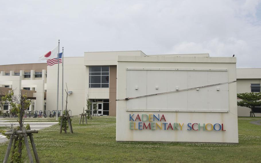 Exterior view of Kadena Elementary School, a modern beige concrete building with large windows and geometric architecture. The school name is displayed in colorful letters on a concrete wall sign in the foreground. Japanese and American flags fly on flagpoles in front of the building.