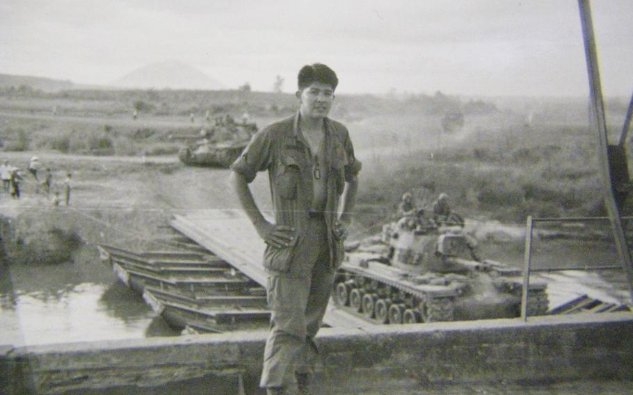 A black-and-white, 1960s-era image of a soldier with his shirt open exposing dog tags standing in front of a bridge as a tank travels across it.
