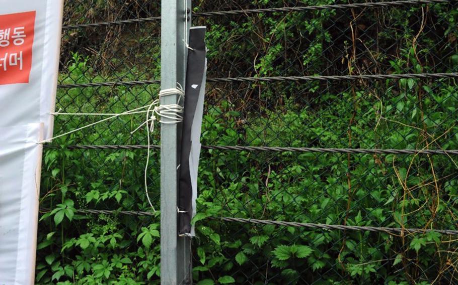 A damaged wire fence is seen with string tied around a post connected to a banner, with dense green vegetation behind it.