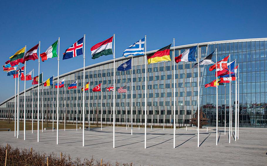 NATO headquarters building with member nations’ flags displayed on white poles in the foreground. The modern glass-faced structure features the NATO flag prominently centered among national flags against a clear blue sky.