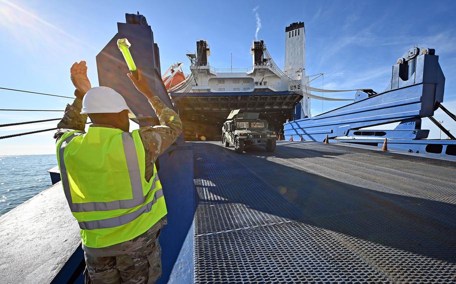 A soldier in a yellow vest raises his arms to give directions as a vehicle is moved off a ship.