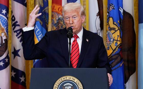 Donald Trump gestures with his hand as he speaks into a microphone while standing at a podium in front of a row of state flags.