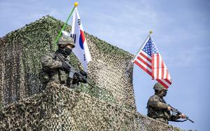 Two soldier in combat gear and holding machine guns stand at the top of a guard tower under U.S. and South Korean flags.