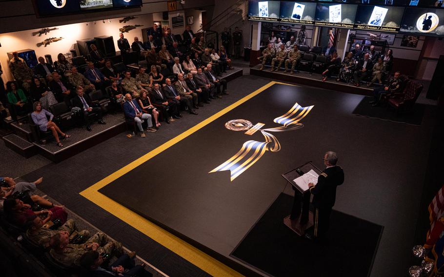 A man in the bottom right stands at a podium, as seen from above, before a crowd and stage bearing an illustration of a medal.