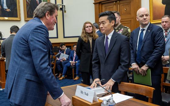 Two men in suits speak while standing on opposite sides of a witness table in a congressional hearing room.