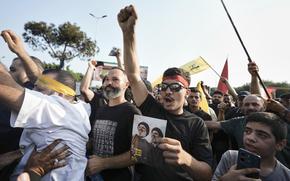 Men raise their fists and cheer or shout outdoors, some holding flags or pictures.
