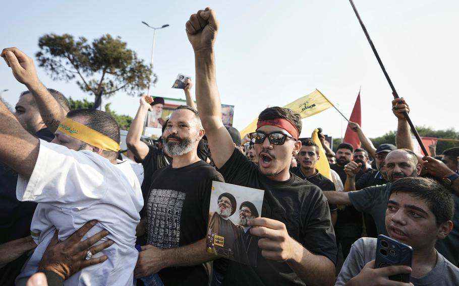 Men raise their fists and cheer or shout outdoors, some holding flags or pictures.