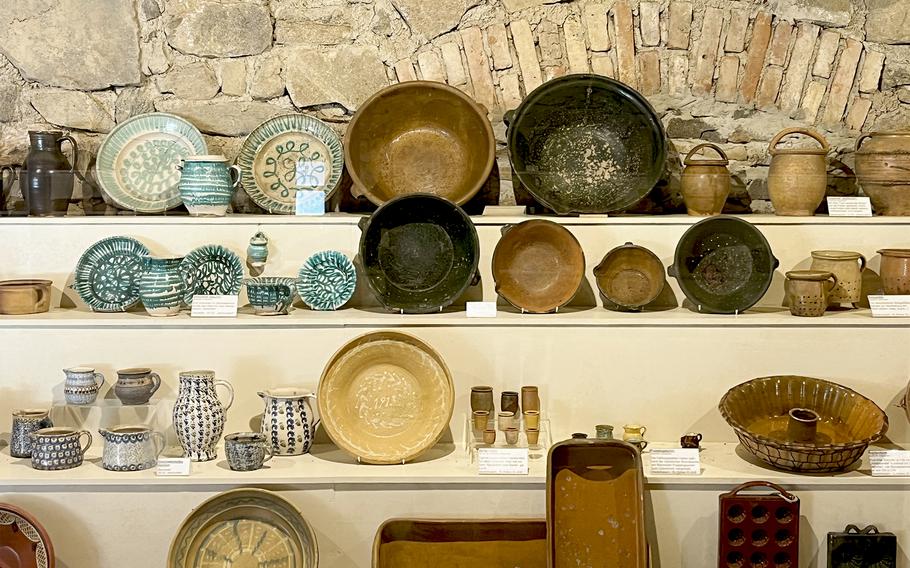 Display shelves in a museum showing traditional pottery and ceramics including decorated plates, bowls, jugs and vessels in various sizes, colors and patterns against a stone wall.