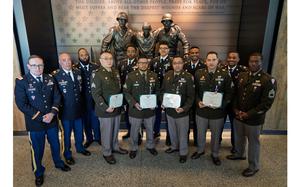 Soldiers in green and blue uniforms stand before a statue, some of them holding certificates.