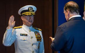 A Navy officer in white dress uniform and hat takes an oath of office with his right hand raised.