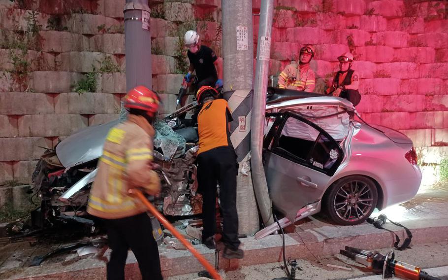 Emergency responders and rescue workers in safety helmets and high-visibility gear work at the scene of a car accident. A severely damaged silver sedan is wedged between a utility pole and a stone block wall. The vehicle shows extensive front-end damage and deployed airbags. Part of the scene is illuminated by emergency lighting, creating a reddish glow on some of the stone walls. 
