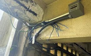 Interior view of a room showing exposed ceiling with peeling paint, electrical conduit and multiple blue wires running along the walls with frayed ends next to a small white electrical box.