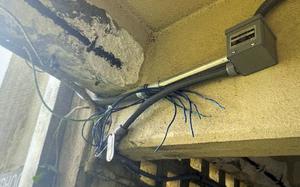 Interior view of a room showing exposed ceiling with peeling paint, electrical conduit and multiple blue wires running along the walls with frayed ends next to a small white electrical box.