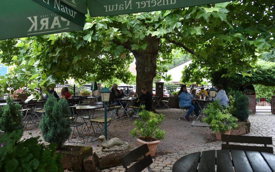  The outdoor dining terrace of a restaurant under large leafy trees, with green umbrellas and dark metal tables and chairs. Diners are seated at various tables enjoying their meals in the dappled shade, creating a pleasant garden restaurant atmosphere.