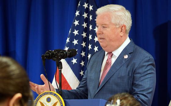 A man in a blue plaid suit with a red tie speaks at a podium bearing the seal of the United States Embassy. He stands in front of a U.S. flag and blue curtain, addressing an audience with microphones positioned in front of him.