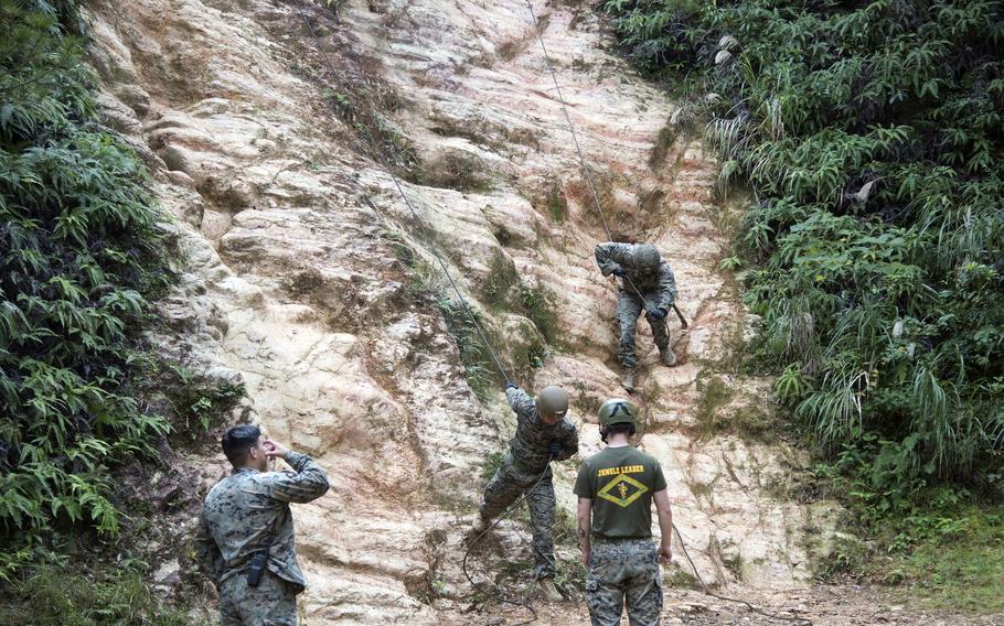 Two Marines stand at the base of a cliff as two others descend it on ropes.
