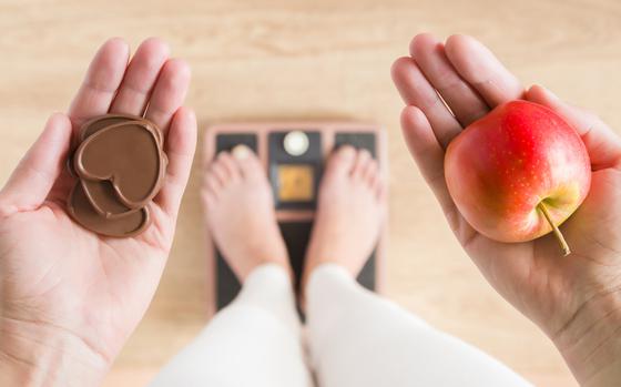 Woman standing on scales and holding apple and chocolate hearts. New start for healthy nutrition, body slimming, weight loss. Cares about body. Dilemma between fruits or sweets. Decision concept.