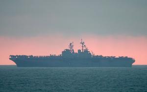 A U.S. Navy amphibious assault ship is silhouetted against a dramatic sunset sky with pink and orange hues. 