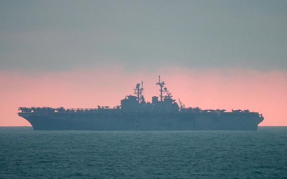 A U.S. Navy amphibious assault ship is silhouetted against a dramatic sunset sky with pink and orange hues. 